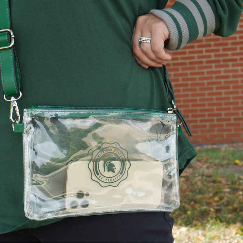 A woman in a green shirt carrying a clear rectangular pouch with green zipper. Decorative Michigan State University circle design printed on front. D-ring attachments on each side with a green long crossbody strap. 