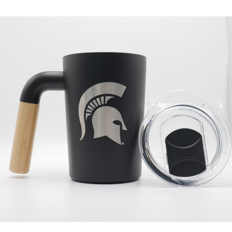 A black, 12 fluid ounce stainless steel coffee mug tumbler with a wooden handle. Engraved into the side of the tumbler in silver stainless steel is a Michigan State spartan helmet logo. The closable clear lid is displayed to the right of the tumbler. 