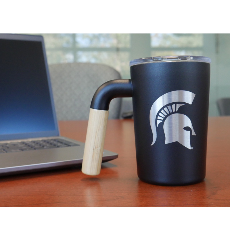 A black, 12 fluid ounce stainless steel coffee mug tumbler with a wooden handle. Engraved into the side of the tumbler in silver stainless steel is a Michigan State spartan helmet logo. The tumbler is sitting on a conference able with a silver laptop computer sitting behind it. 