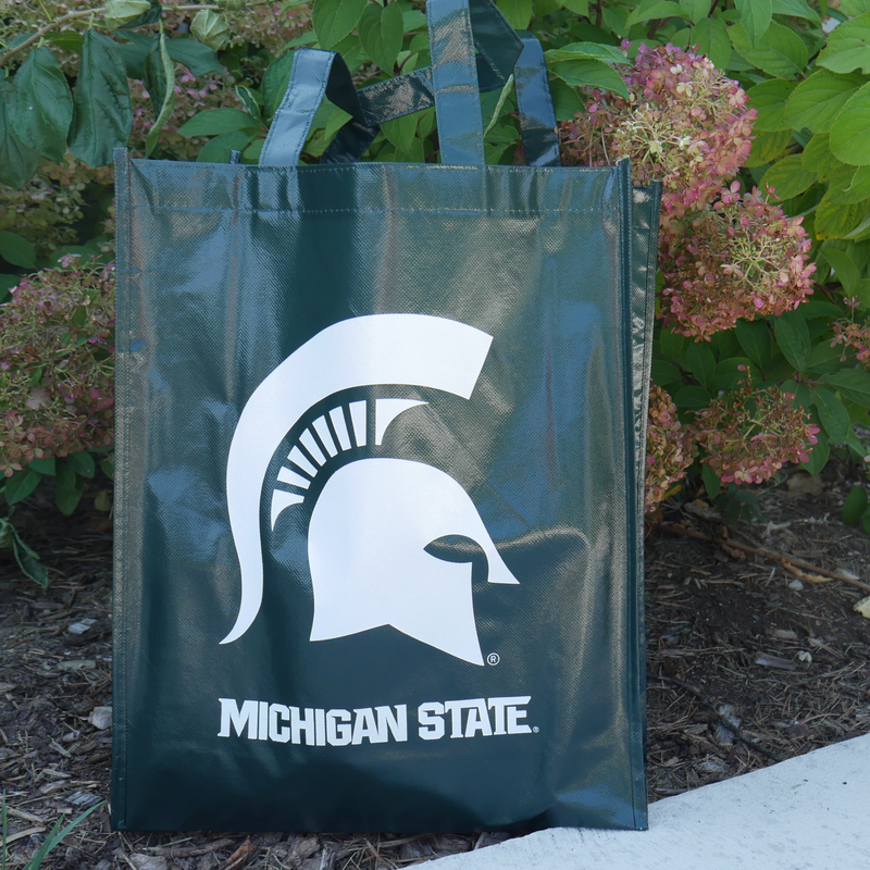 A dark green re-usable laminated tote bag. On the side of the bag read "Michigan State" in white with a large, white MSU spartan helmet logo above the text. The bag is sitting in front of a flower bush. 