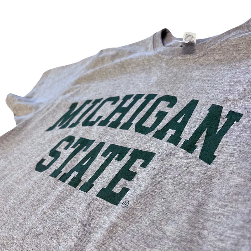 Close up of the chest area of a heather gray t-shirt. Forest green block letters read "Michigan State" across two lines, with "Michigan" slightly arched.