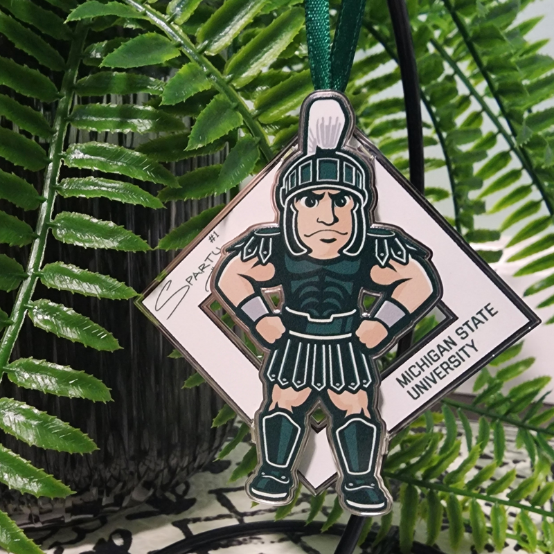 The Sparty collectible on a black ornament stand in front of a potted fern. The collectible is an illustration of MSU’s Sparty mascot sporting a smirk with his hands on his hips. A white diamond behind Sparty displays his "Sparty number 1" signature and athletic block letters that read Michigan State University. A green ribbon is affixed to the top of the collectible.