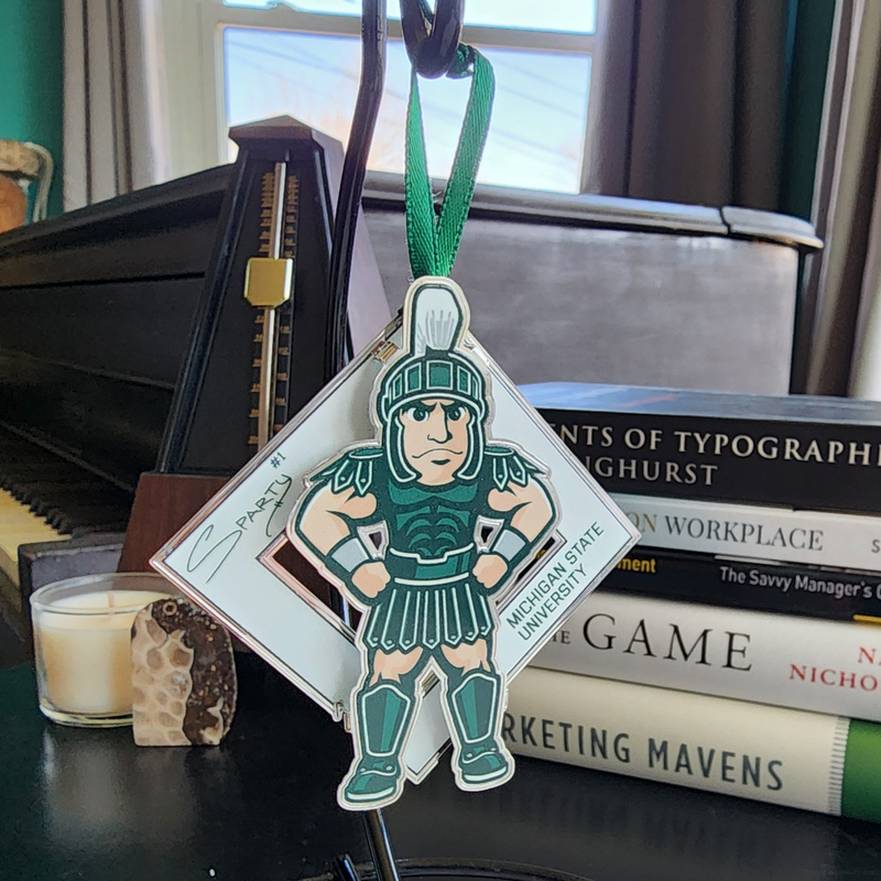 The Sparty collectible on a black ornament stand in front of a piano, stack of books, candles, and a Petoskey stone. The collectible is an illustration of MSU’s Sparty mascot sporting a smirk with his hands on his hips. A white diamond behind Sparty displays his "Sparty number 1" signature and athletic block letters that read Michigan State University. A green ribbon is affixed to the top of the collectible.