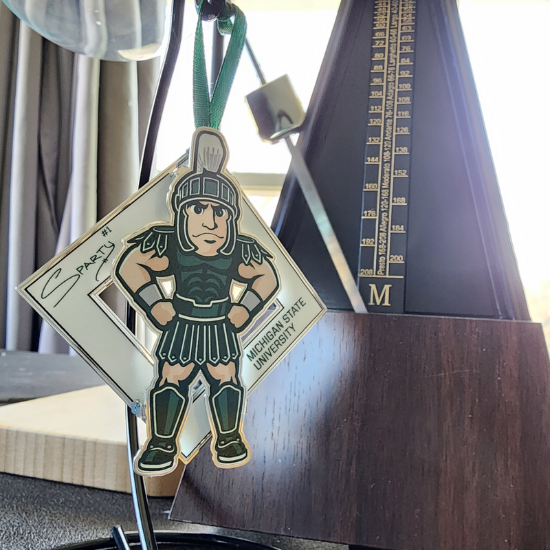 The Sparty collectible on a black ornament stand in front of a window, a metronome, and lamp. The collectible is an illustration of MSU’s Sparty mascot sporting a smirk with his hands on his hips. A white diamond behind Sparty displays his "Sparty number 1" signature and athletic block letters that read Michigan State University. A green ribbon is affixed to the top of the collectible.