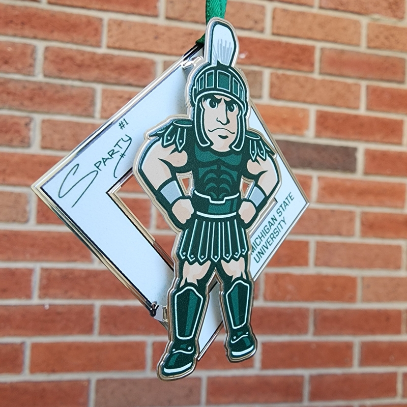 The Sparty collectible in front of a brick wall. The collectible is an illustration of MSU’s Sparty mascot sporting a smirk with his hands on his hips. A white diamond behind Sparty displays his "Sparty number 1" signature and athletic block letters that read Michigan State University. A green ribbon is affixed to the top of the collectible.