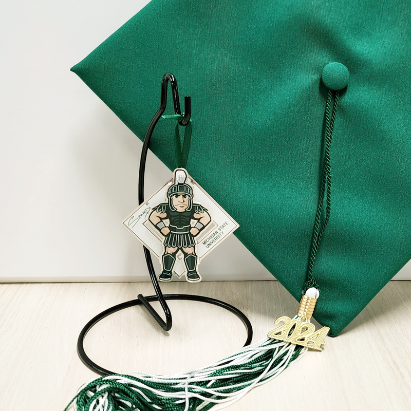 The Sparty collectible on a black ornament stand next to an MSU graduation cap with a 2024 commemorative tassel. The collectible is an illustration of MSU’s Sparty mascot sporting a smirk with his hands on his hips. A white diamond behind Sparty displays his "Sparty number 1" signature and athletic block letters that read Michigan State University. A green ribbon is affixed to the top of the collectible.