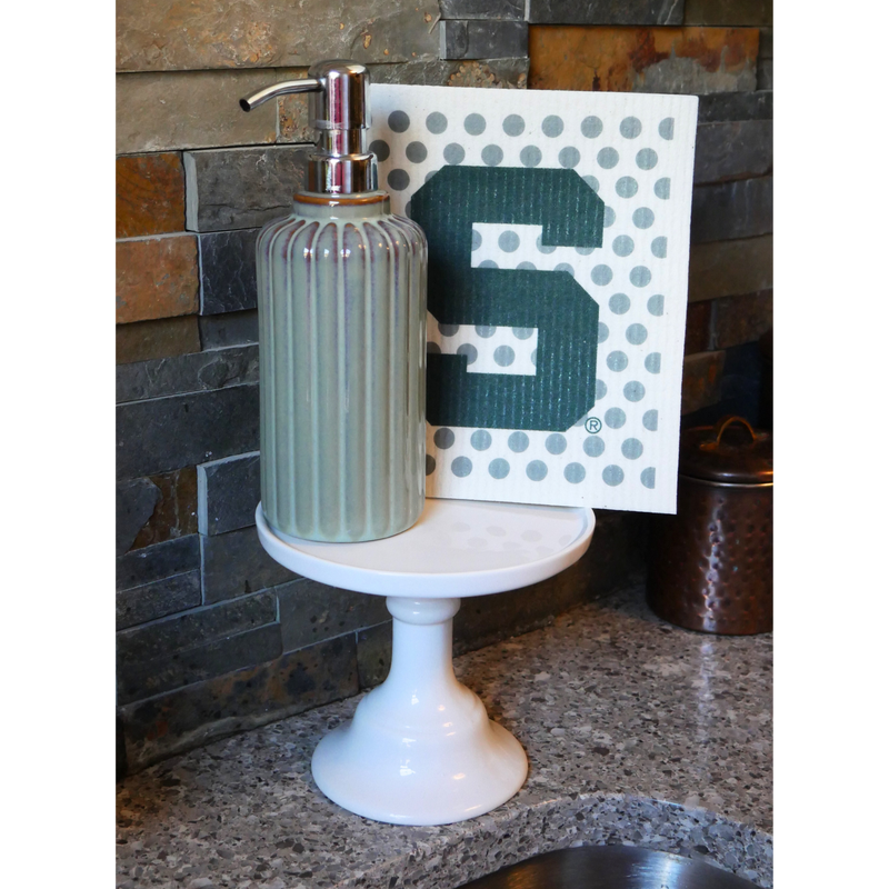 Sitting on a white, small display pedestal next to an olive green soap dispenser is a cream re-usable absorbent square dish cloth with a gray polka-dot pattern. In front of the pattern in the center of the dish cloth is a dark green Michigan State block S logo. 