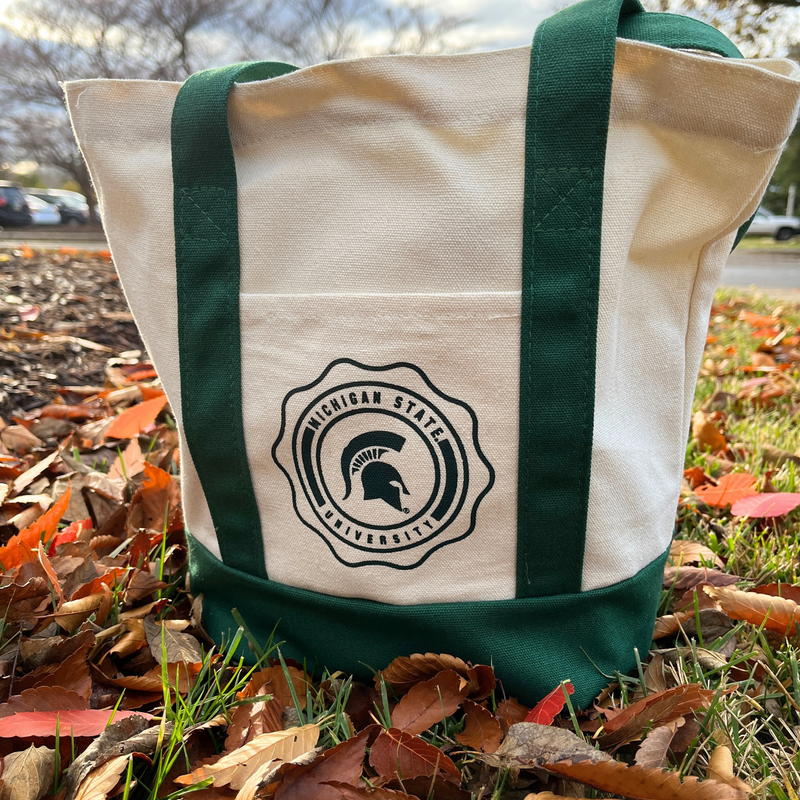 A cream colored canvas tote bag with dark green handles and a dark green bottom. In the middle of the bag is a circular emblem with the words "Michigan State University" around the border. In the middle of the emblem is a green MSU spartan helmet logo. 