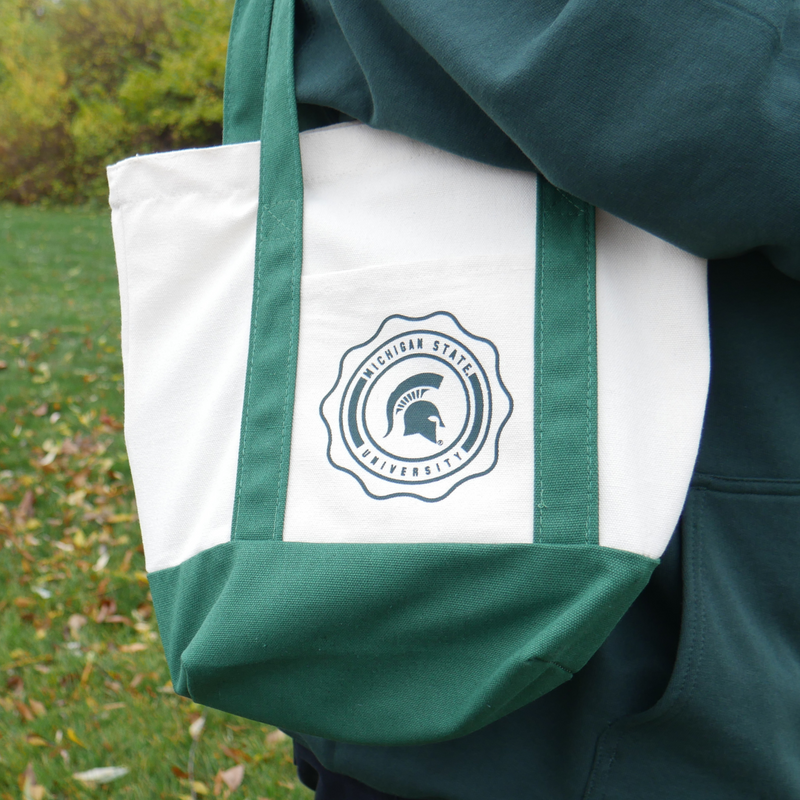 Close up of a woman carrying a cream colored canvas tote bag with dark green handles and a dark green bottom. In the middle of the bag is a circular emblem with the words "Michigan State University" around the border. In the middle of the emblem is a green MSU spartan helmet logo. 