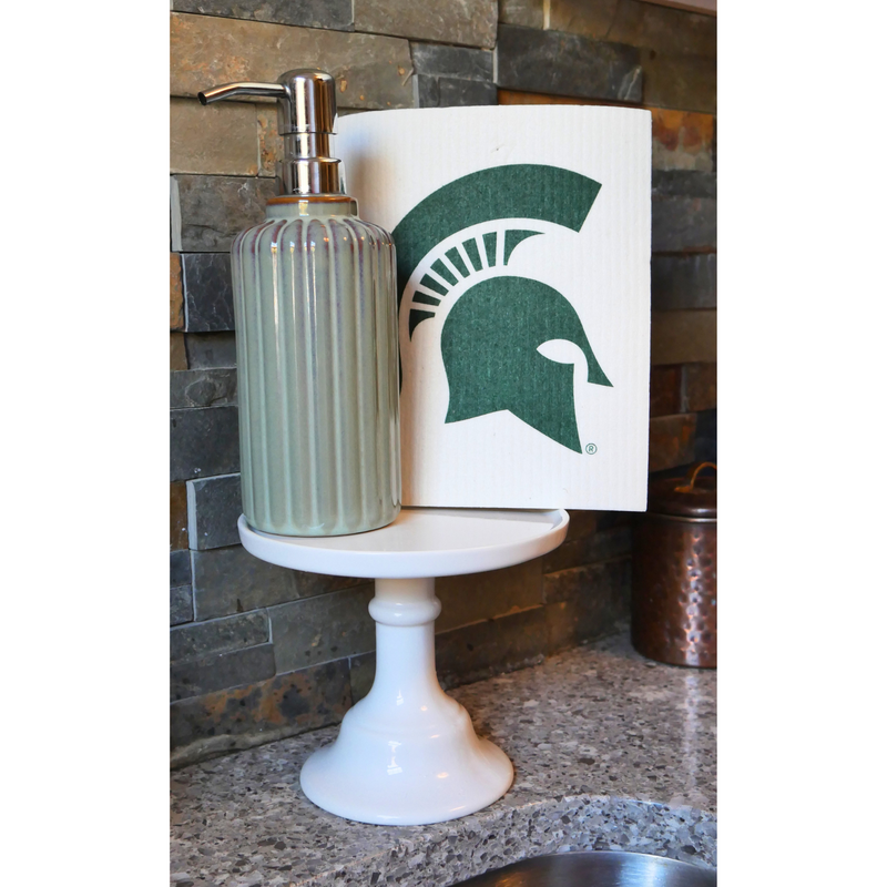 Sitting on a white display pedestal next to an olive green soap dispenser is a cream re-usable absorbent square dish cloth with a dark green Michigan State spartan helmet logo in the center. 