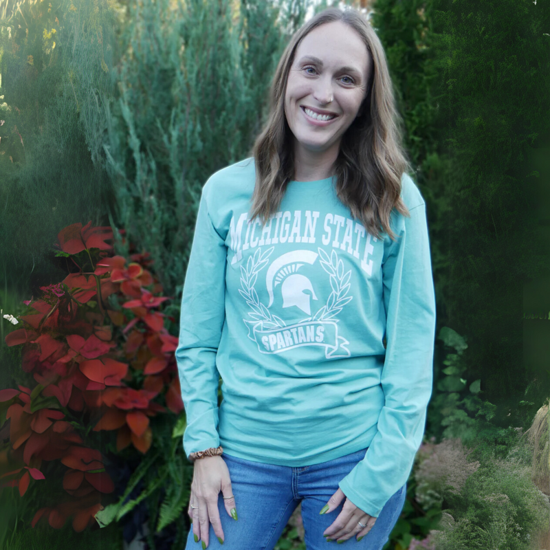 Woman modeling t-shirt outdoors. A mint green long sleeve t-shirt with Michigan State Spartans front center design in white. A wreath surrounds a white MSU spartan helmet logo. 