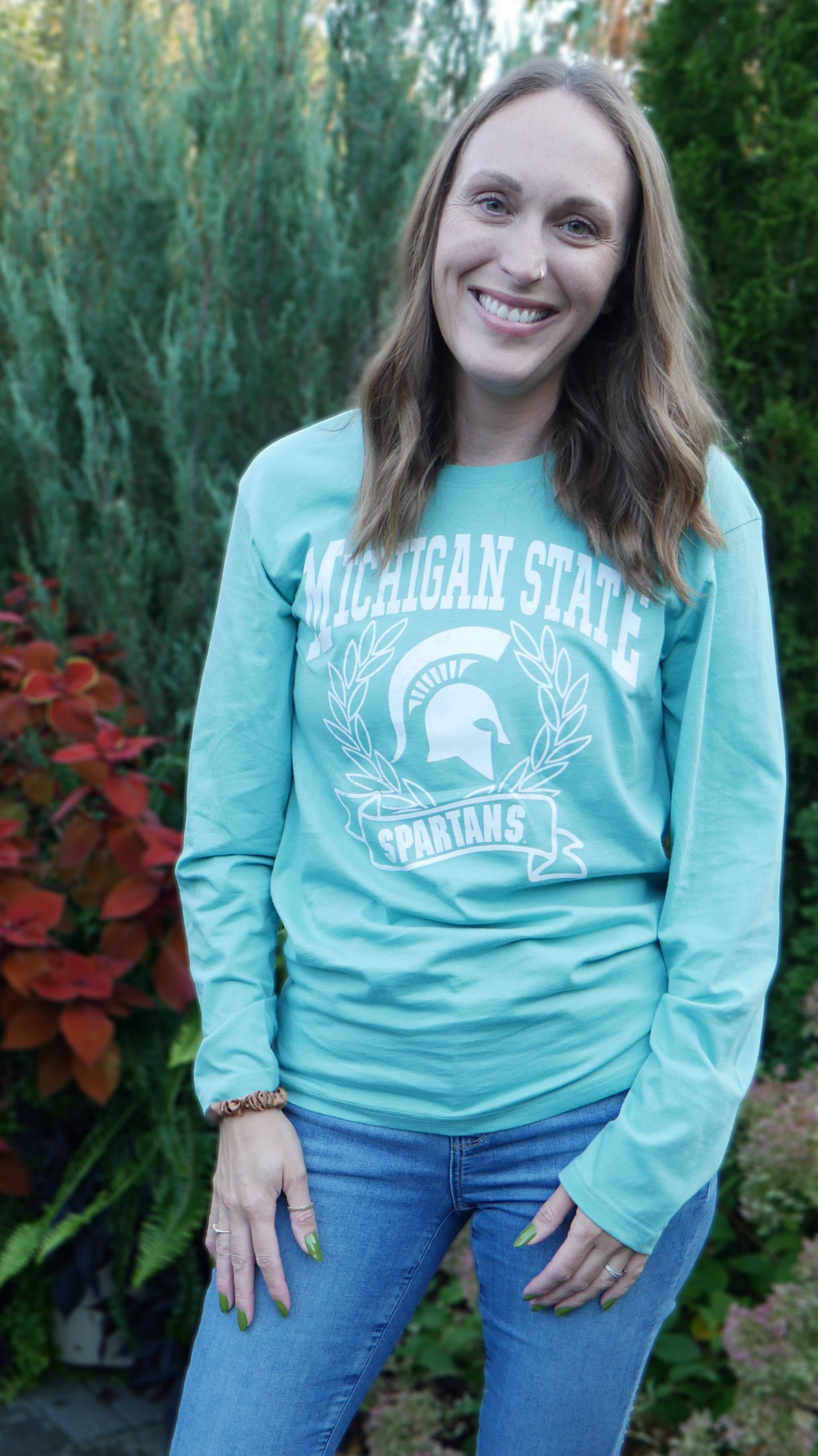 Woman modeling t-shirt outdoors. A mint green long sleeve t-shirt with Michigan State Spartans front center design in white. A wreath surrounds a white MSU spartan helmet logo. 