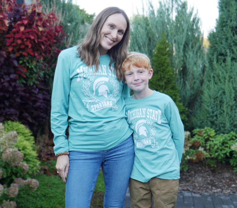 Woman and child modeling t-shirt outdoors. A mint green long sleeve t-shirt with Michigan State Spartans front center design in white. A wreath surrounds a white MSU spartan helmet logo. Adult and youth.