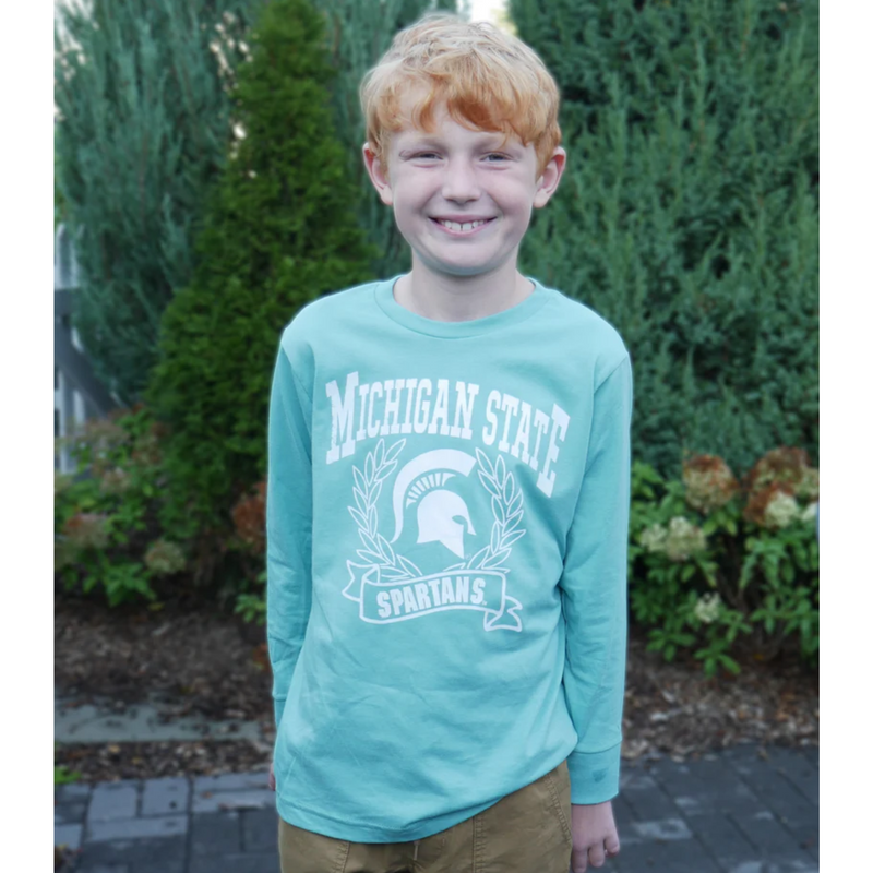 Child modeling t-shirt outdoors. A mint green long sleeve t-shirt with Michigan State Spartans front center design in white. A wreath surrounds a white MSU spartan helmet logo. 