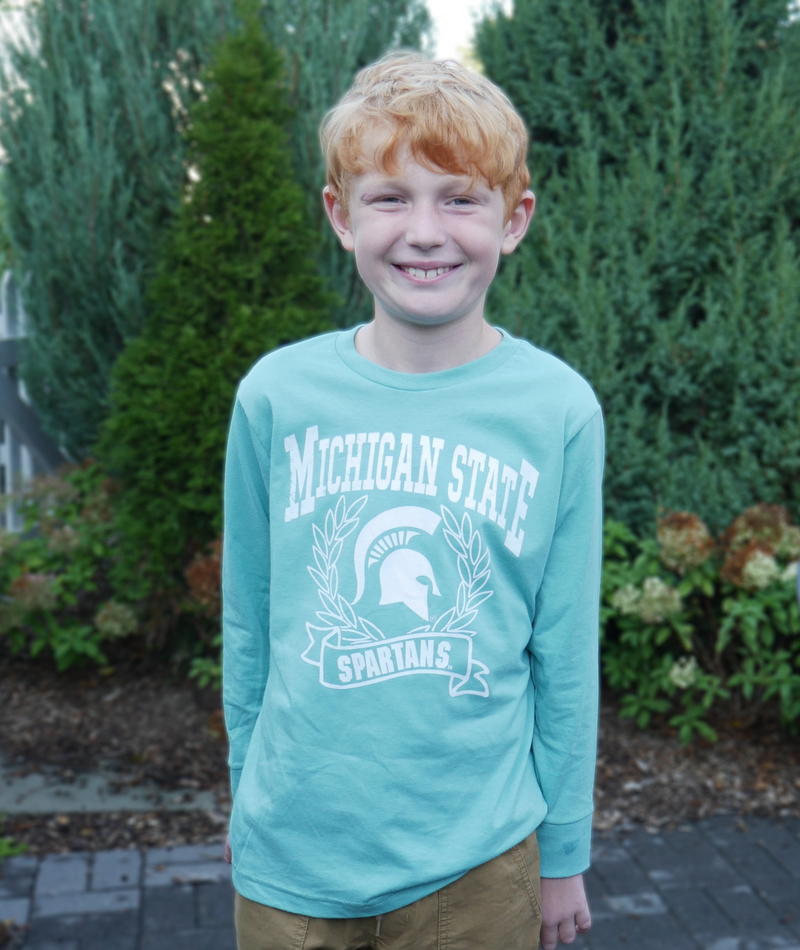 Child modeling t-shirt outdoors. A mint green long sleeve t-shirt with Michigan State Spartans front center design in white. A wreath surrounds a white MSU spartan helmet logo. 