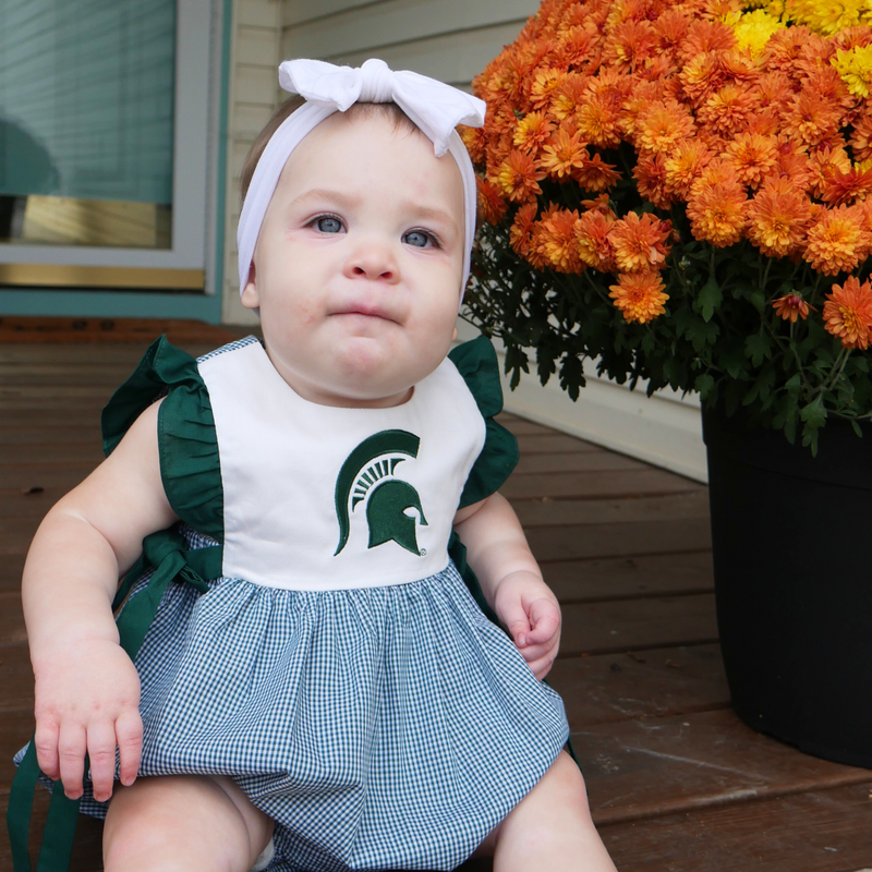 A child wearing a sleeveless baby jumper next to a large bouquet of orange flowers. The green and white plaid bottom has a bubble effect in shape. The top is white with dark green ruffle trim ending in bows at the hip. A dark green Spartan helmet is printed on the center chest.