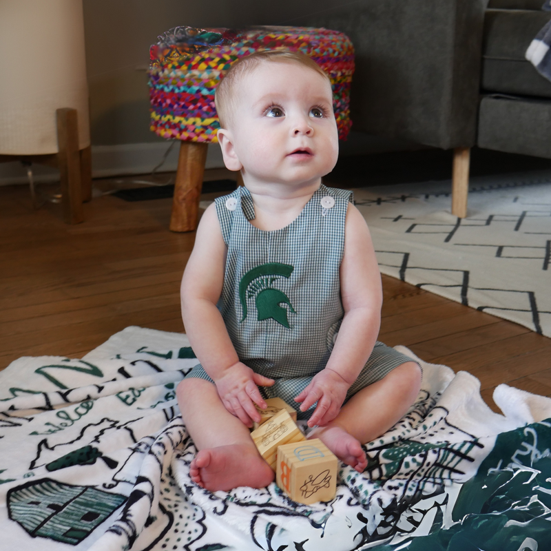 A toddler sitting on a white patterned blanket playing with toy wooden blocks. The toddler is wearing the MSU embroidered infant jumper. 