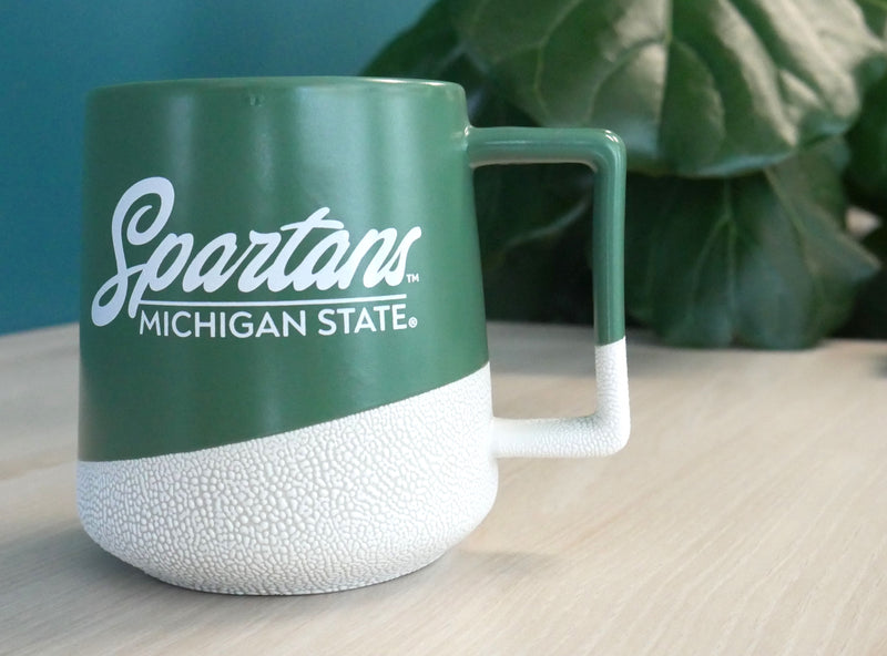 A green and ceramic printed coffee mug with "Spartans: Michigan State" written in white on the side in cursive and all caps print. 