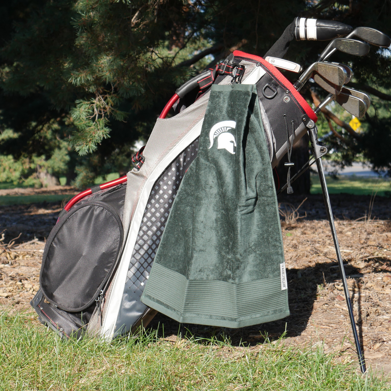 Attached to the side of a golf bag with golf clubs is a dark green golf towel with a black clip at the top to attach to golf bags. On the towel is a white MSU spartan helmet logo. 