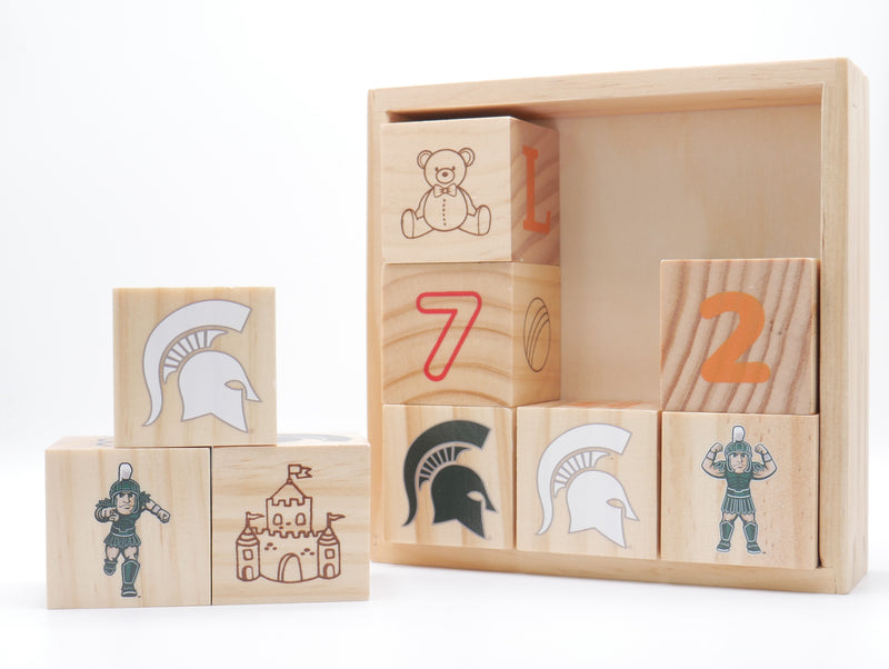 A set of nine wooden play blocks. Six of the blocks are stacked inside a wood storage box showing various MSU-related icons and numbers. A smaller stack of three blocks is on the left side of the box showing Sparty, the MSU spartan helmet logo, and a drawing of a castle. 