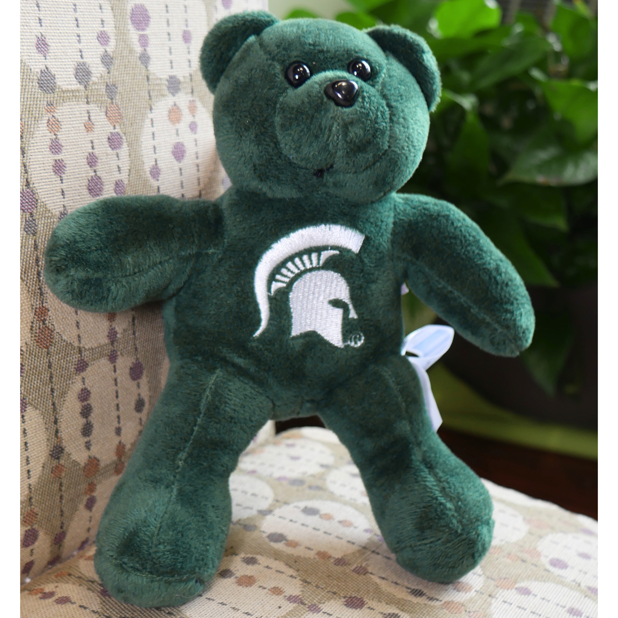 A green plush teddy bear sitting on a brown and tan couch. On the chest of the bear is a white MSU spartan helmet logo. 