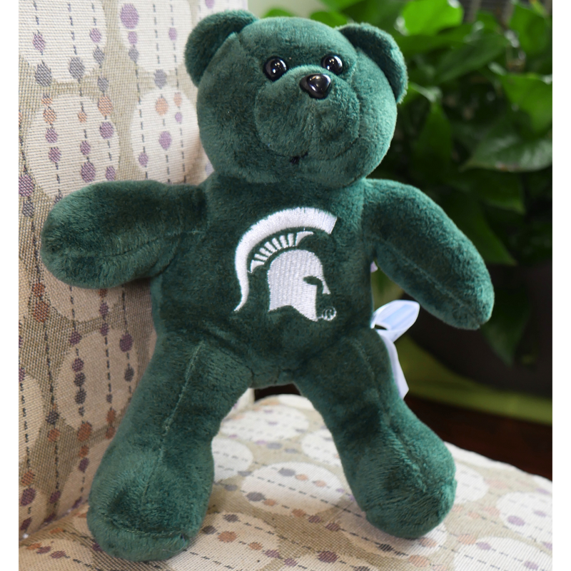 A green plush teddy bear sitting on a brown and tan couch. On the chest of the bear is a white MSU spartan helmet logo. 