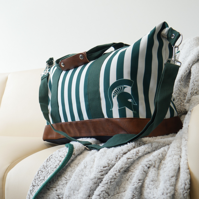 Laying on top of a silver blanket and a yellow couch is a green and white vertical striped canvas tote bag with a brown leather bottom and strap handles. The bag also has a green crossbody strap. In the lower right corner of the tote bag is a green MSU spartan helmet logo. 