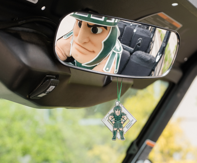 Sparty's reflection in the rearview mirror of a golf cart. Hanging from the mirror is the brass 2024 Sparty Signature collectible, which measures 2.75 inches in height and features Sparty with his hands on his hips in front of a white cut-out diamond shape.