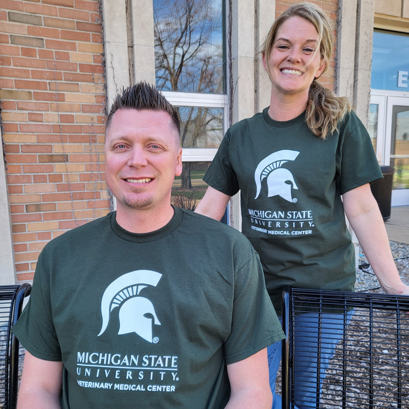 A man and a woman wearing a green short-sleeve t-shirt. Across the center chest is a large white Spartan helmet centered above the Michigan State wordmark logo. Beneath that is large block text reading “Veterinary Medical Center.”