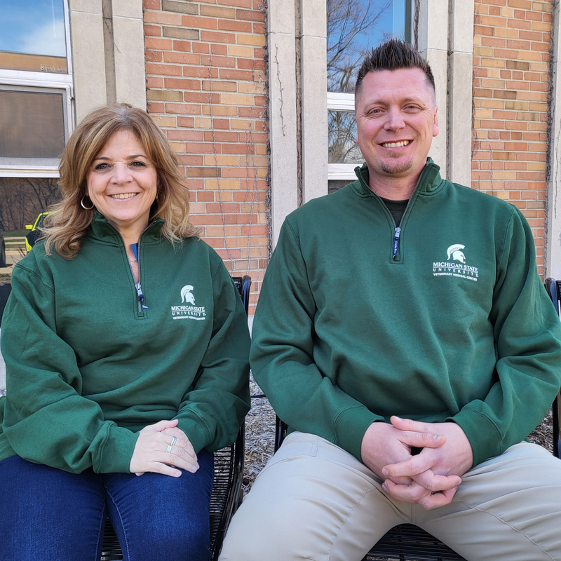 A man and a woman wearing a dark green, quarter zip sweatshirt. The upper left chest is embroidered with a Veterinary Medical Center logo in white.