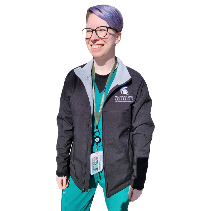 A woman in green scrubs wearing a black full-zip jacket. The upper left chest is embroidered with a Veterinary Medical Center logo in white.