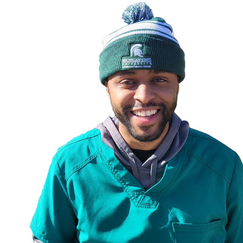 A man in green scrubs sitting on a bench wearing a green and white striped beanie. The beanie is embroidered with a Veterinary Medical Center logo in white.