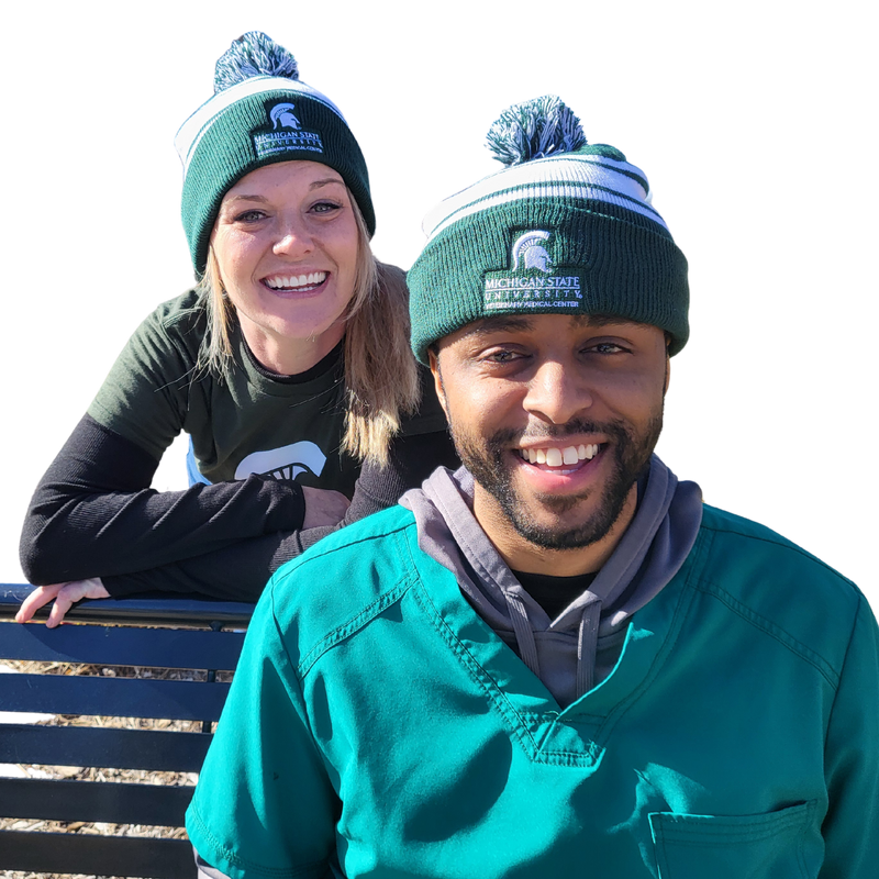 A man in green scrubs sitting on a bench wearing a green and white striped beanie. The beanie is embroidered with a Veterinary Medical Center logo in white. Behind and to the left of the man is a woman leaning on the bench wearing the same beanie.