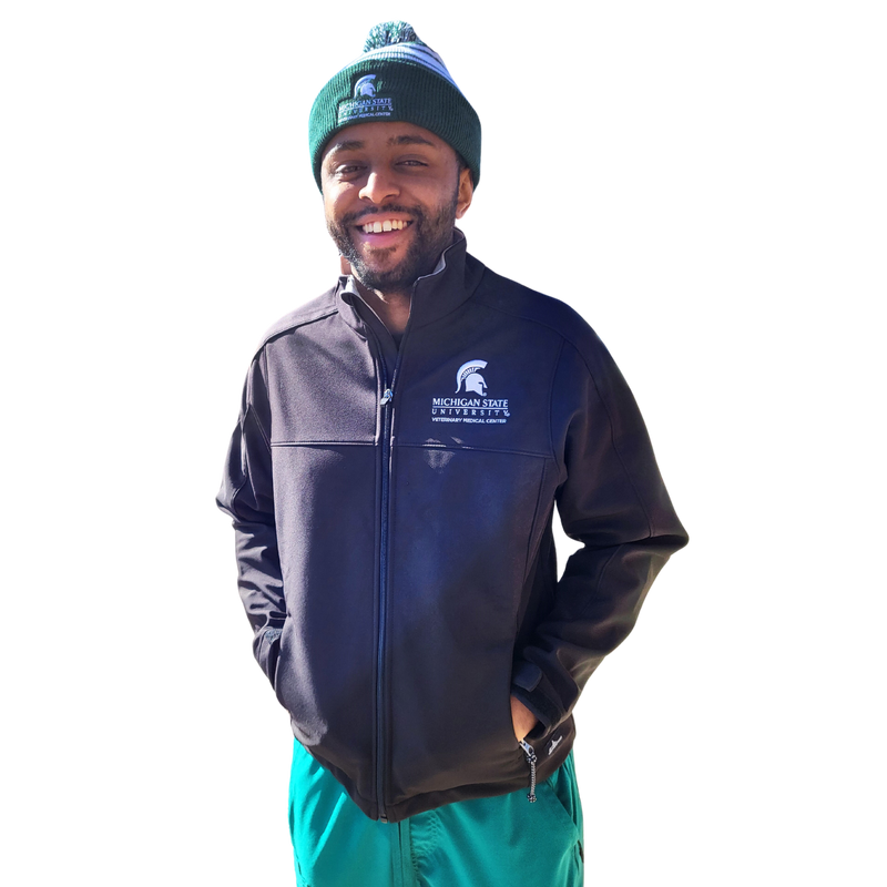 A man in green scrubs and a green beanie wearing a black full-zip jacket. The upper left chest is embroidered with a Veterinary Medical Center logo in white. 