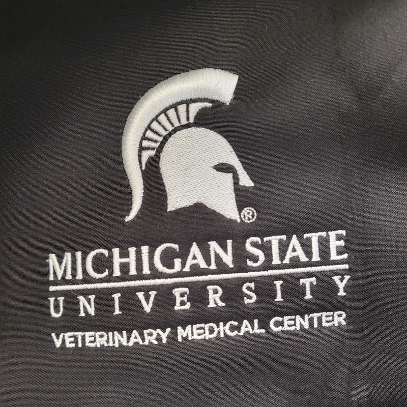 Embroidered in white, a large Spartan helmet is centered over the Michigan State University wordmark logo, with block letters reading “Veterinary Medical Center” beneath. 