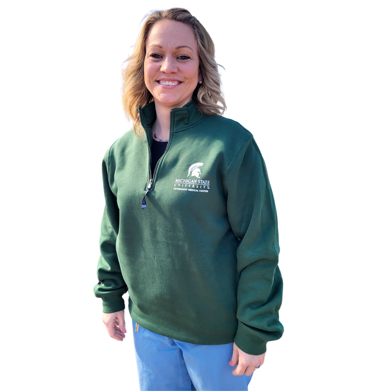 A woman in blue scrubs wearing a dark green, quarter zip sweatshirt. The upper left chest is embroidered with a Veterinary Medical Center logo in white.
