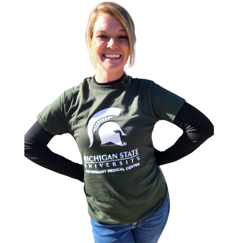 A woman in blue jeans wearing a green short-sleeve t-shirt. Across the center chest is a large white Spartan helmet centered above the Michigan State wordmark logo. Beneath that is large block text reading “Veterinary Medical Center.”