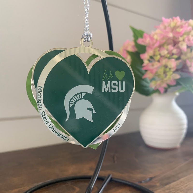 Green heart ornament with a white Spartan helmet and We [heart] MSU. Interlaced with a silver circle reading Michigan State University and 2023. Ornament hanging from a black hanger sitting on a wooden shelf with a white shiplap background and a white vase with pink flowers.