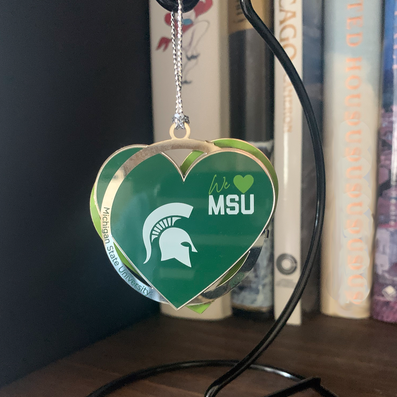 Green heart ornament with a white Spartan helmet and We [heart] MSU. Interlaced with a silver circle reading Michigan State University and 2023. Ornament hanging from a black hanger sitting on a wooden shelf with a black background and several books.