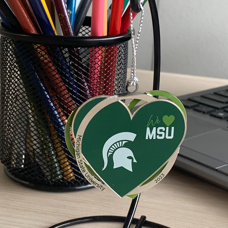 We Love MSU ornament - a green heart with a silver circle reading Michigan State University and 2023 and a lime green heart outline back. Ornament is hanging from a holder and sitting on a desk with colored pencils and a laptop in the background.