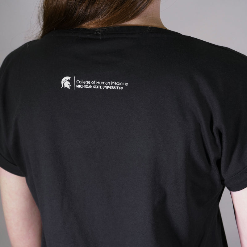 Back of a woman wearing a black short sleeve t-shirt with the College of Human Medicine signature logo printed just below the neckline.