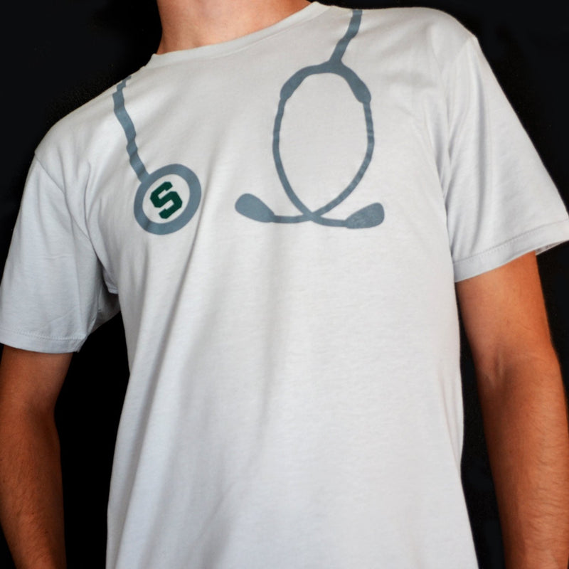 Man wearing a white t-shirt with a graphic of a silver stethoscope around the shoulders. The circle of the stethoscope has a green block S in the center.