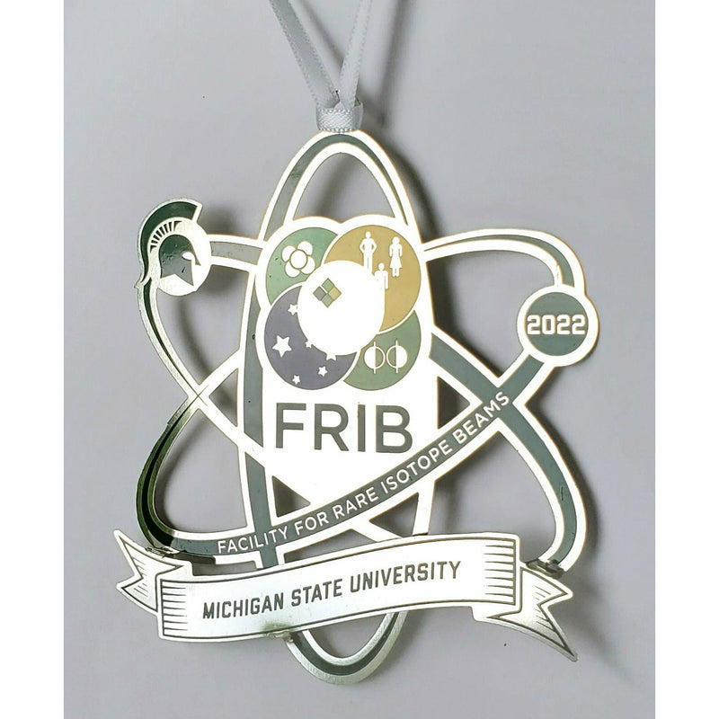 Silver metal ornament on a white ribbon, cut in the shape of an atom. Text reads "FRIB: Facility for Rare Isotope Beams" with an electron reading "2022" and a metal ribbon reading "Michigan State University"