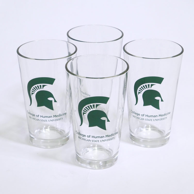 Collection of four clear tapered pint glasses with a large green Spartan helmet printed on the center. Below the helmet, green text reads College of Human Medicine Michigan State University on two lines.