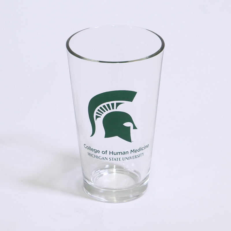 Clear tapered pint glass with a large green Spartan helmet printed on the center. Below the helmet, green text reads College of Human Medicine Michigan State University on two lines.