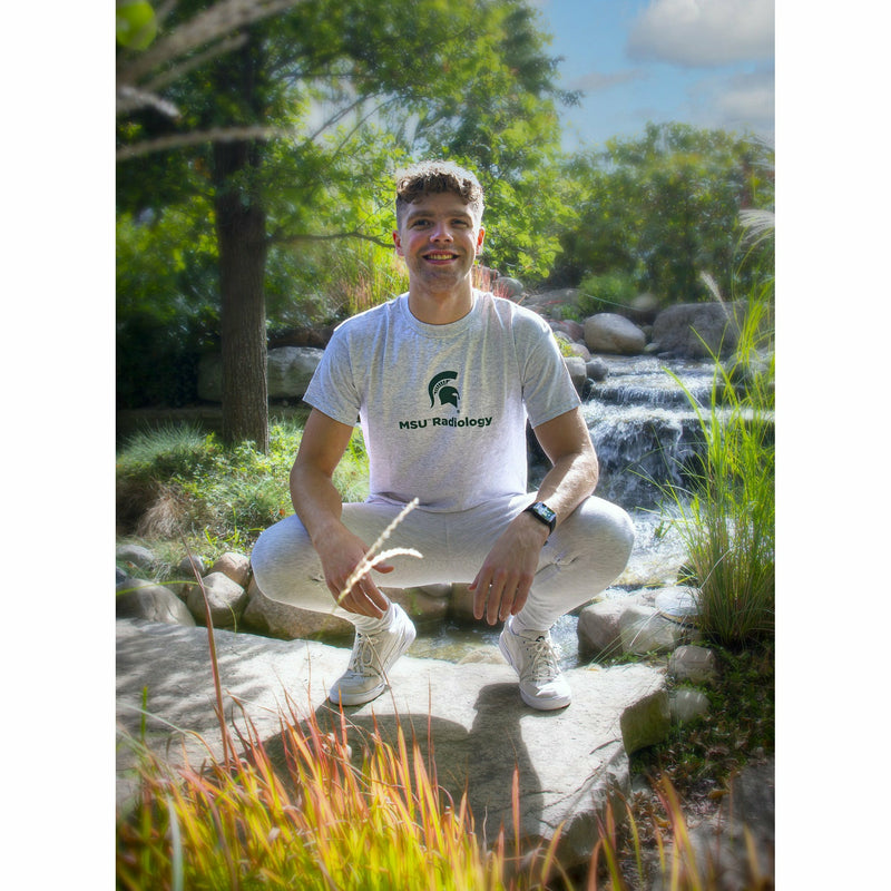 A young man squats in front of a waterfall within the gardens to show the light gray t-shirt.
