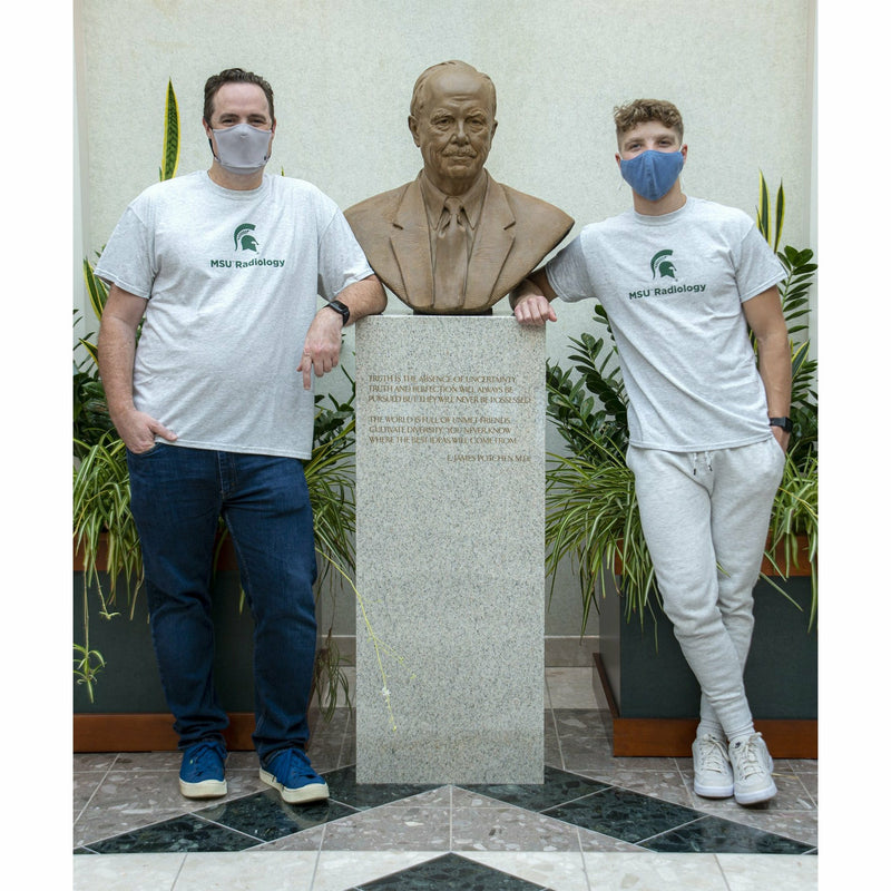 Two men stand next to the bust of Dr. E. James Potchen wearing the light gray t-shirts, showing what the shirt looks like on different body types.