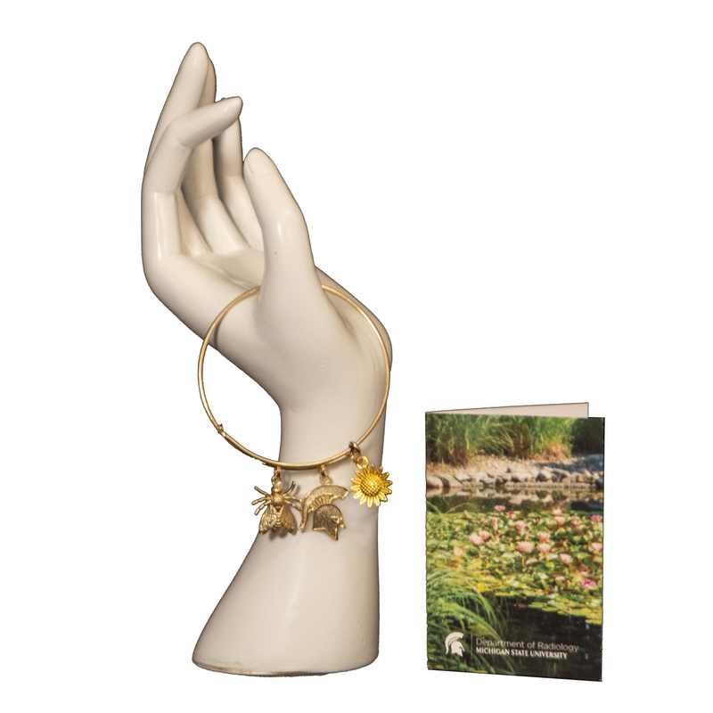 The gold bee and sunflower bracelet on a hand mannequin next to a greeting card featuring a photo of a pond in the gardens.