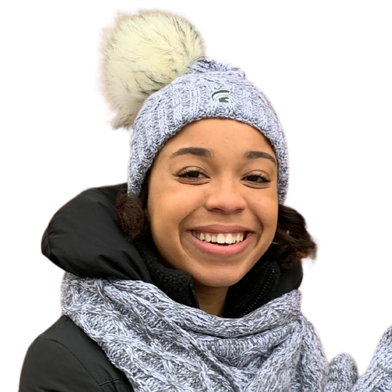 Close-up of a woman wearing a light gray knit beanie with an off-white and gray pom at the top. On the front is an embroidered green Spartan helmet.