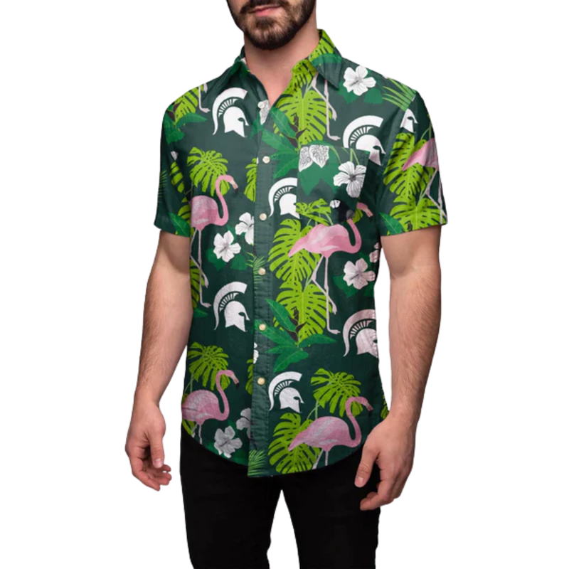 Front view of a man wearing the Michigan State Spartans Floral Button-Up over black slacks. The shirt has a dark green background with pink flamingos, white hibiscus flowers, white Spartan helmets, and a variety of green leaves.